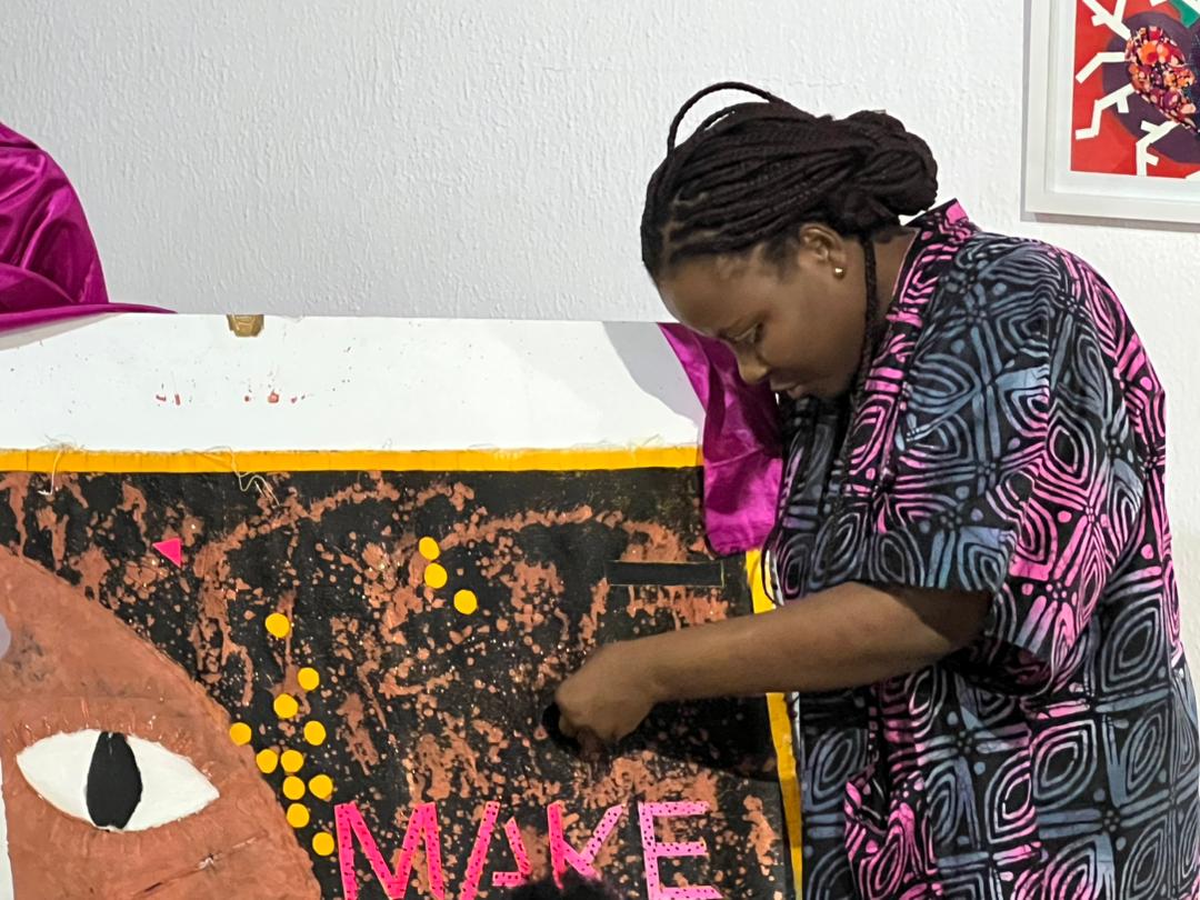 A woman in a pink and blue patterned shirt works on a large mixed-media canvas with an eye, yellow dots and pink lettering