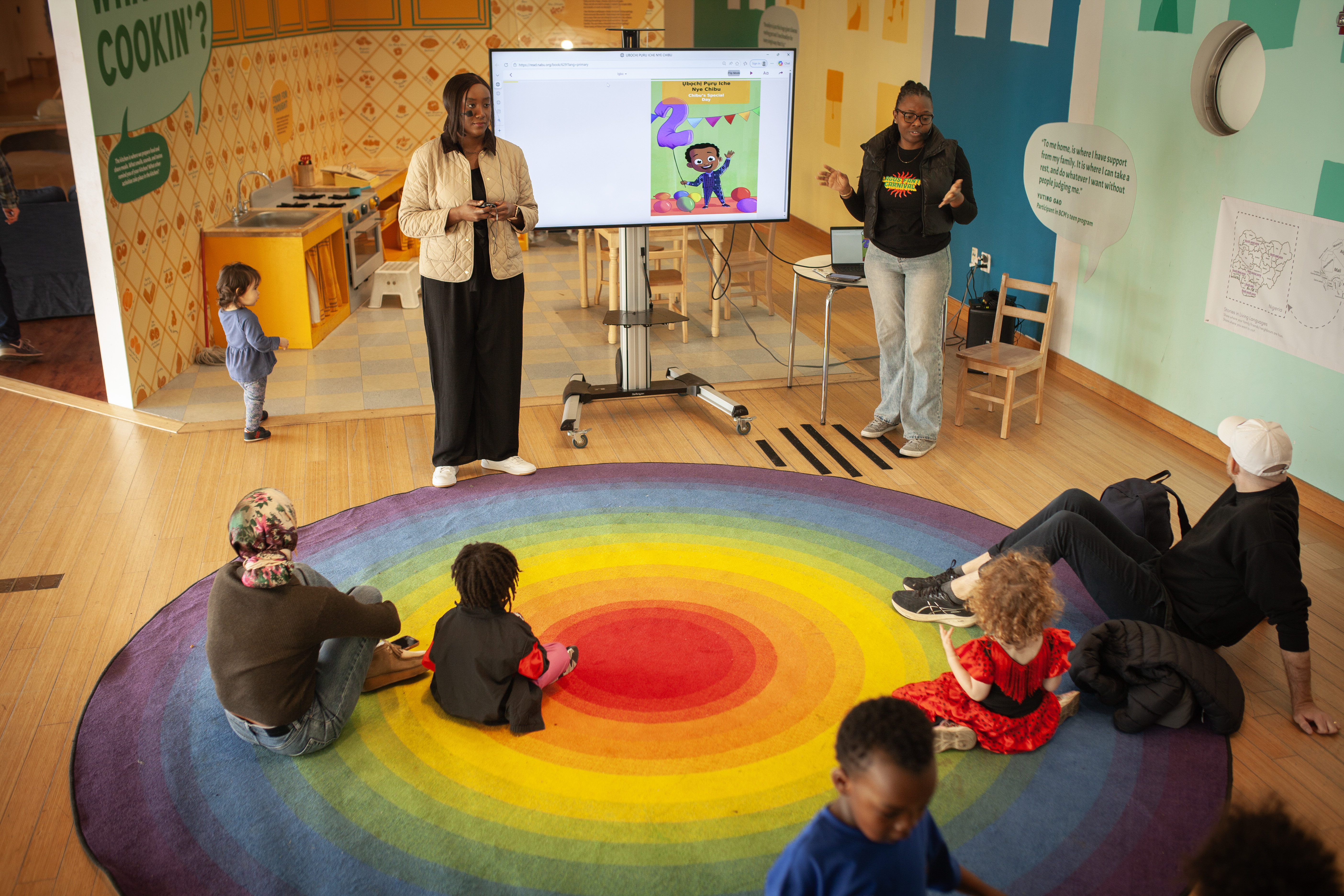 Stories in Living Languages at Brooklyn Children's Museum — presenters beside a screen with participants gathered on a rainbow rug
