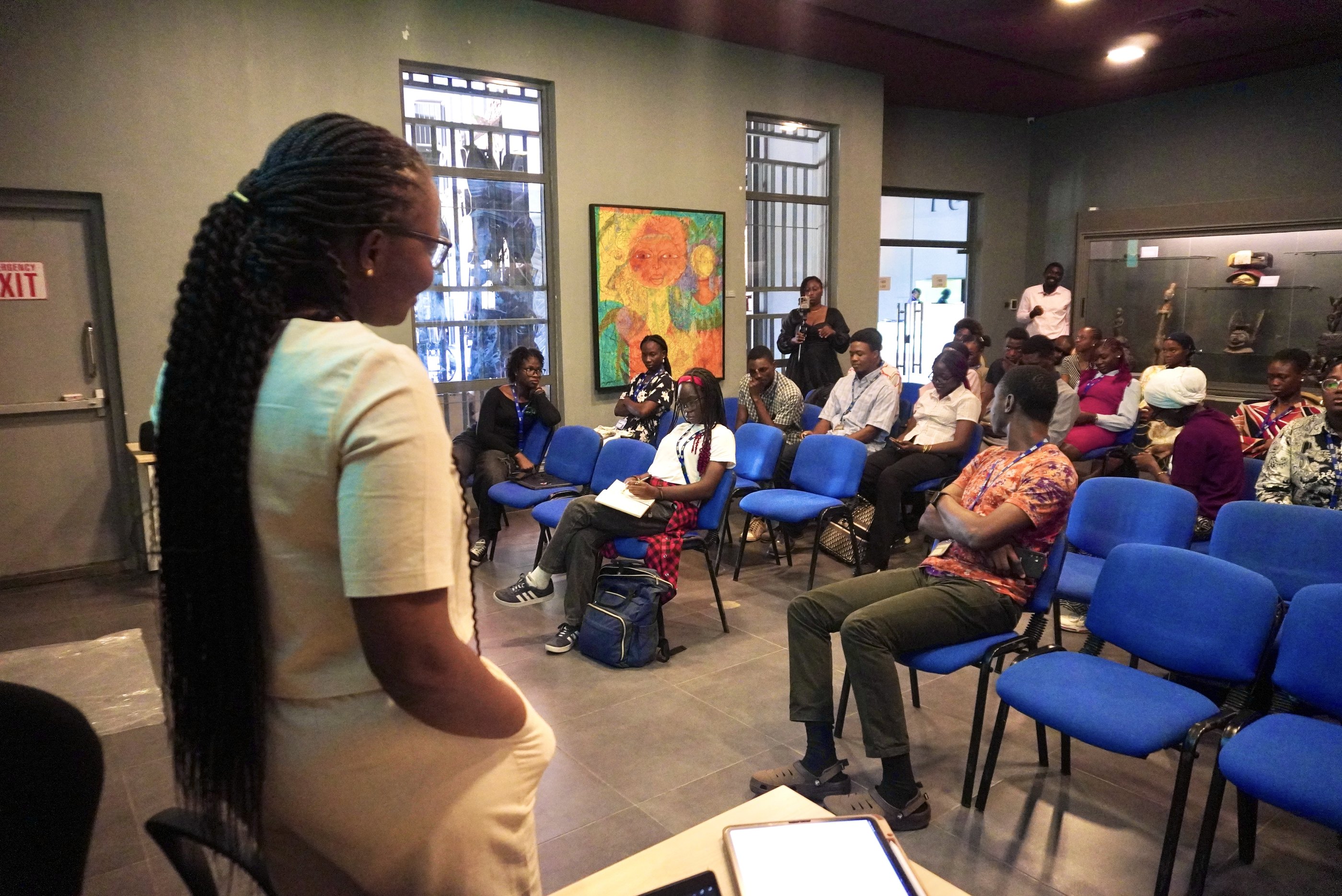 A facilitator addresses participants seated on blue chairs in a museum gallery room