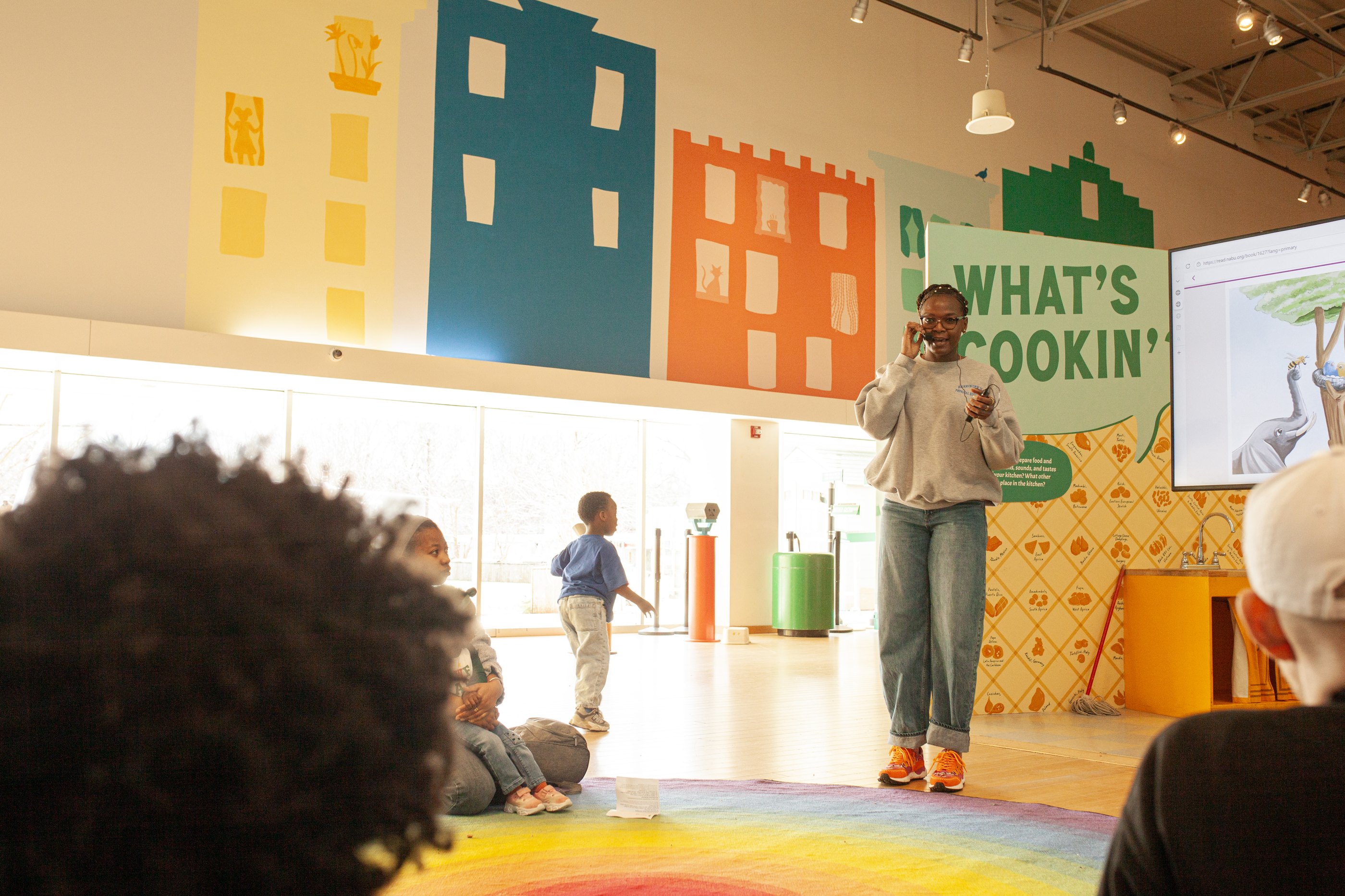 A presenter speaks to seated participants in a mural-painted museum space, with a screen beside her