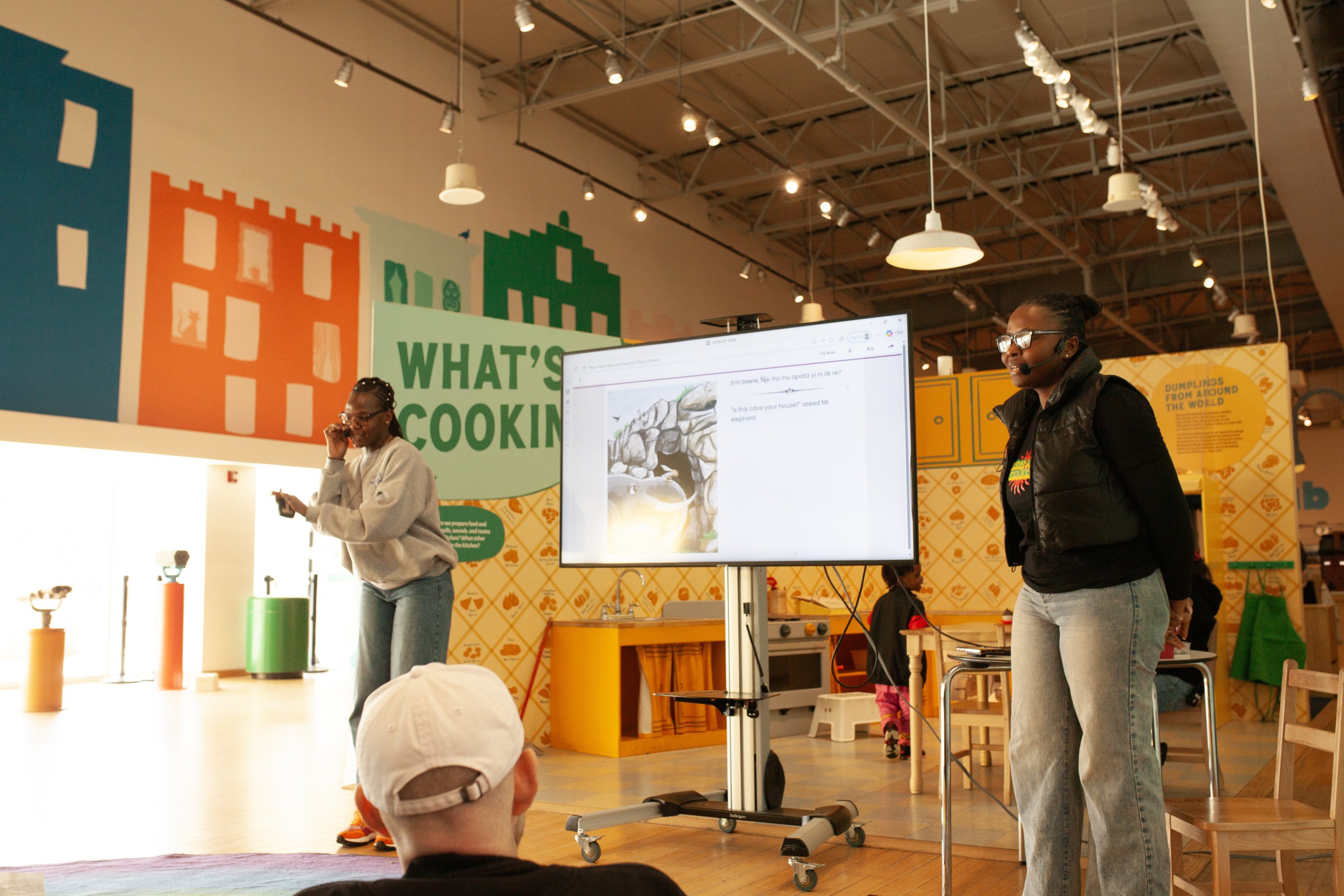 Two presenters stand beside a screen showing an illustrated book, with participants seated in front