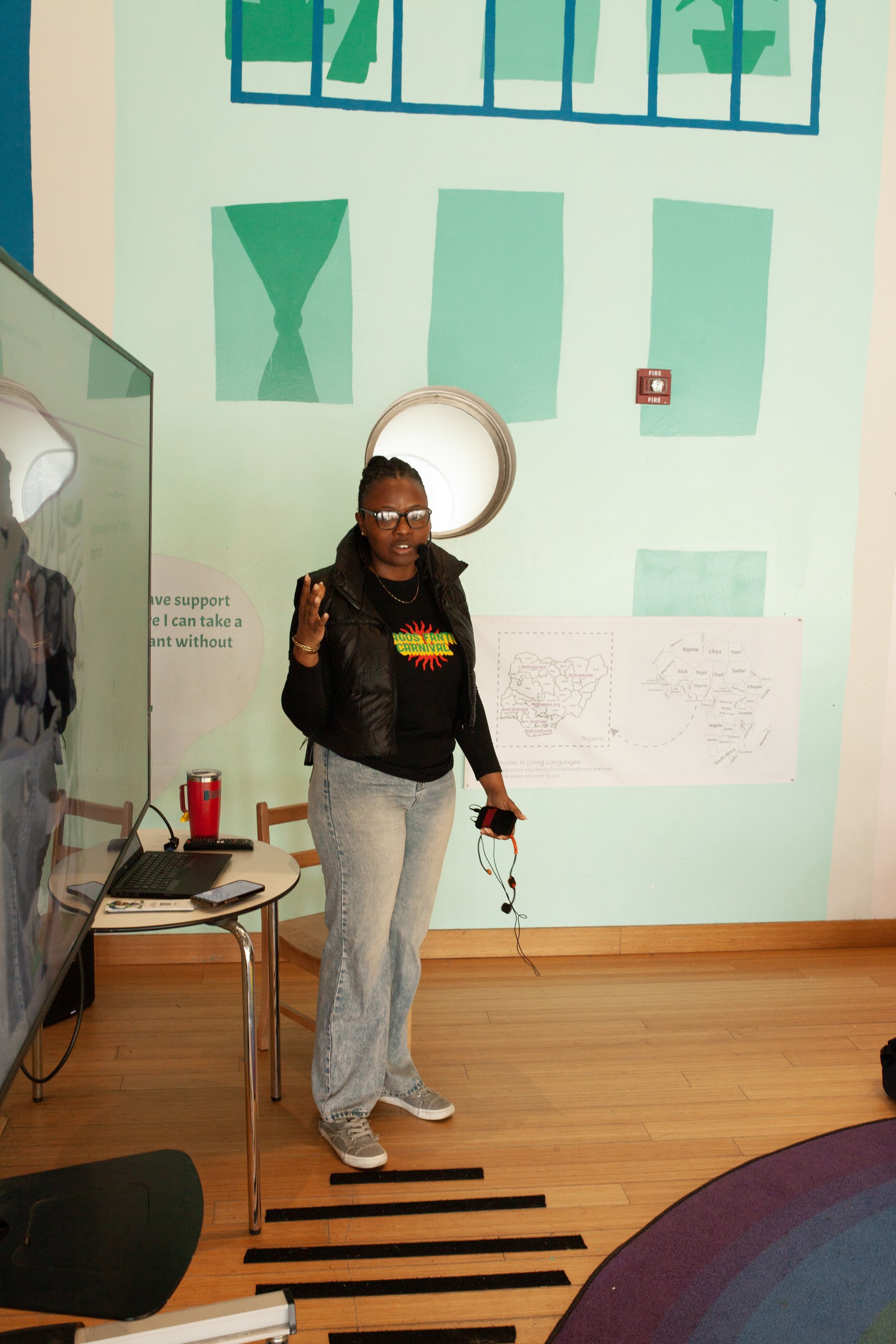 A presenter gestures while speaking, standing before a pastel mural and a wall map